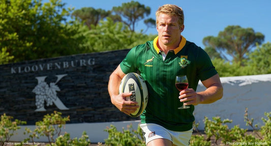 Pieter-Stef Du Toit, wearing his green Springbok jersey stands outdoors in front of a Kloovenburg Wine Estate sign. He is holding a rugby ball in one hand and a glass of red wine in the other, with vineyard greenery and blue sky in the background.
